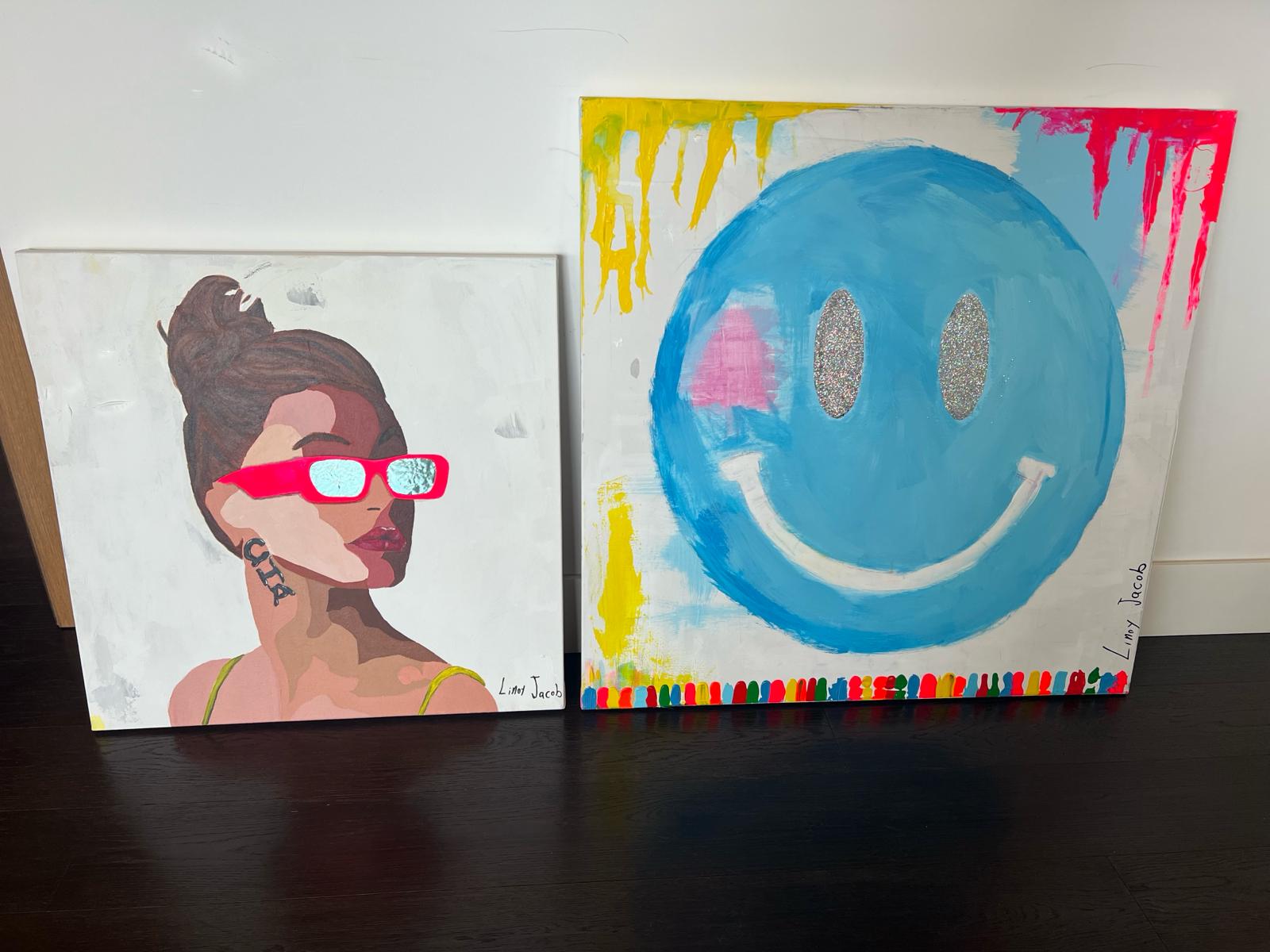 Blue Glitter Smiley painting and Hailey Bieber Pop Art portrait by Linoy Jacob displayed together on a dark wood floor